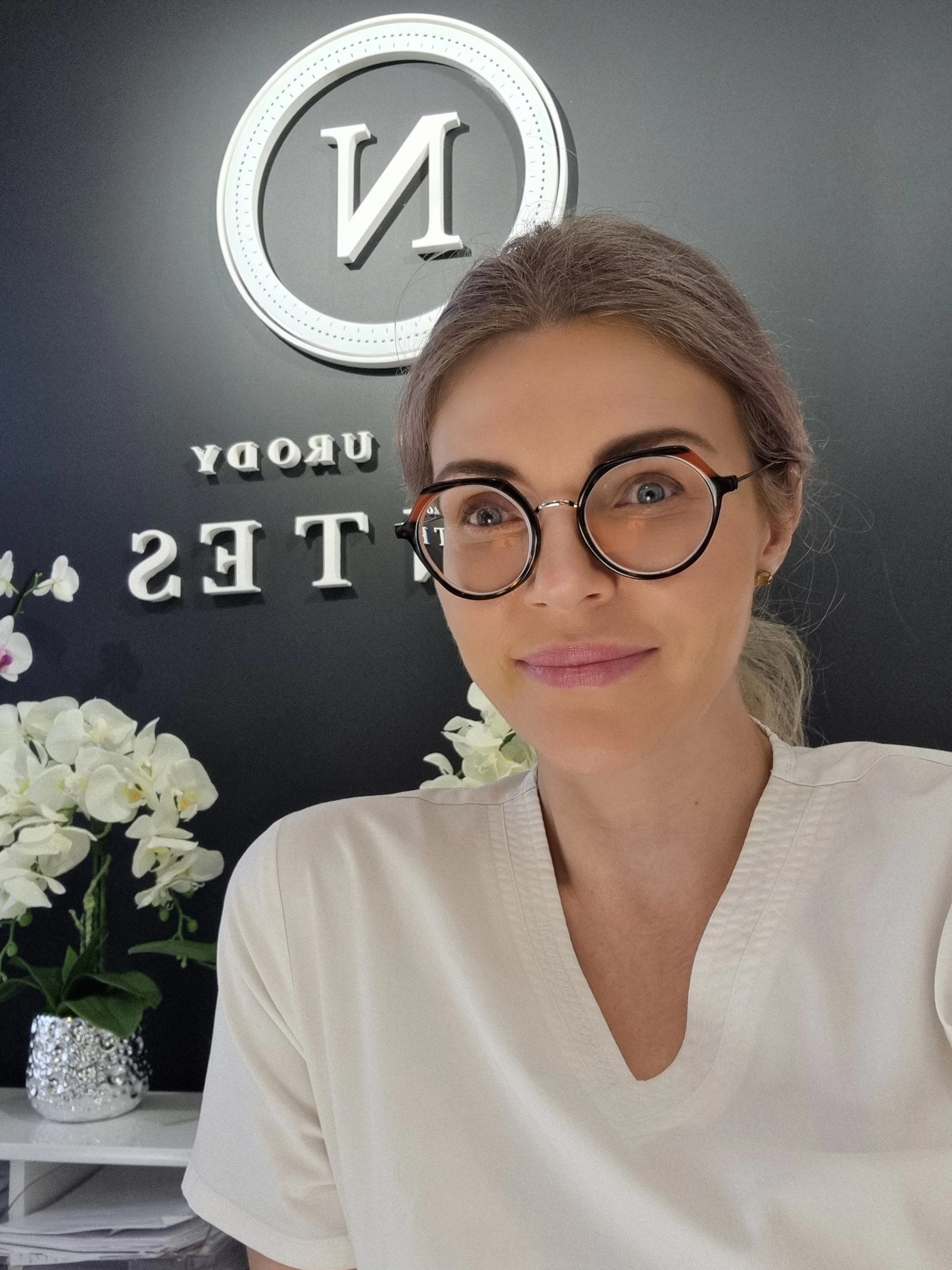 lek. Magdalena Olczyk, uśmiechnięty lekarz medycyny estetycznej z Nantes Med & Beauty w Płocku, w białym uniformie i charakterystycznych okularach, w recepcji gabinetu na tle logo.